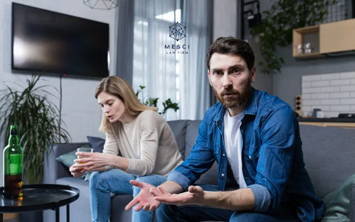 A couple in the process of divorce sitting in their living room looking in different directions; the man looks pensively at the camera while the woman, with a drink in her hand, gazes in another direction.